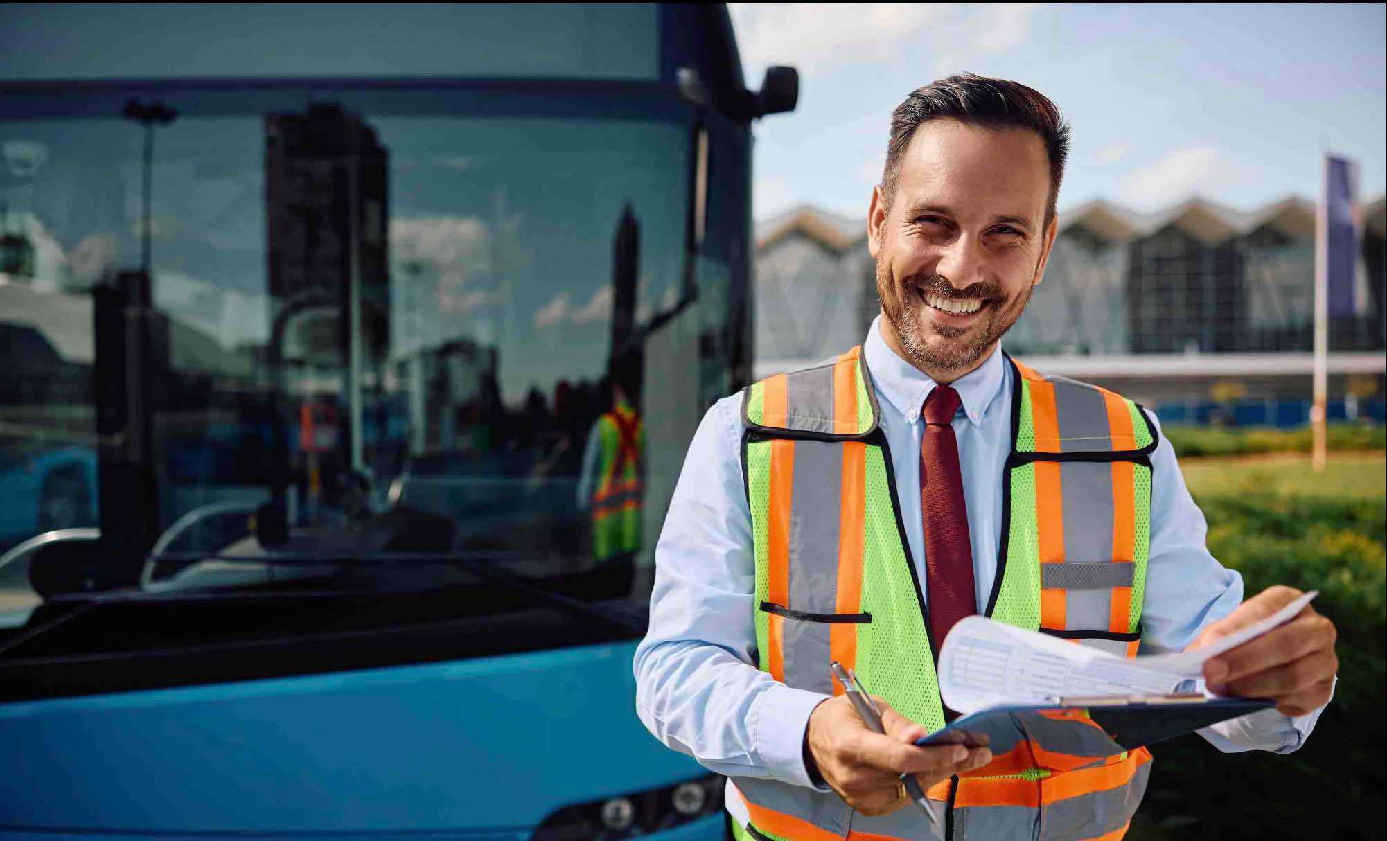 A smiling transit supervisor holding a clipboard in front of a bus